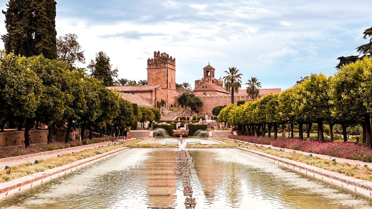UnViaggioPerDue: Una vista panoramica dei giardini dell'alcazar di Cordoba con giochi d'acqua simmetrici e giardini curati che conducono all'antica fortezza sotto un cielo nuvoloso.