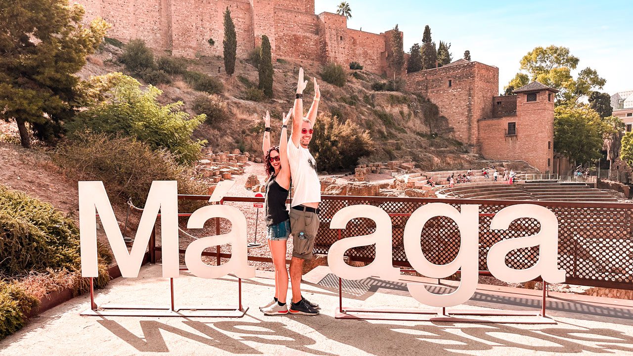 UnViaggioPerDue: Daniele e Flavia dietro un grande cartello "Malaga", con le mura storiche dell'Alcazaba sullo sfondo in una giornata di sole.