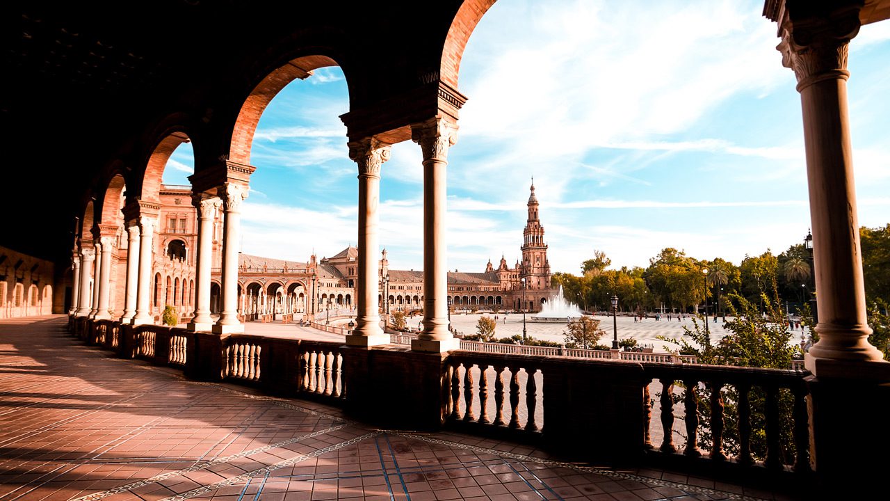 UnViaggioPerDue: Veduta di Piazza di Spagna a Siviglia, con archi lungo un corridoio con intricate lavorazioni di piastrelle, che conduce a un edificio centrale attraverso un canale, sotto un cielo limpido.