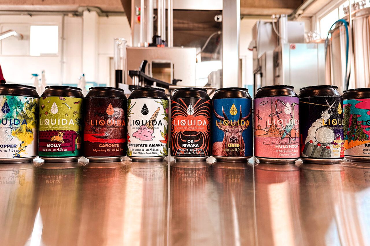 UnViaggioPerDue: Una fila di lattine di birra colorate del Birrificio Artigianale Liquida esposte su una superficie metallica in un birrificio nella provincia di Ferrara. Ciascuna lattina presenta grafica e marchio vivaci e distinti.