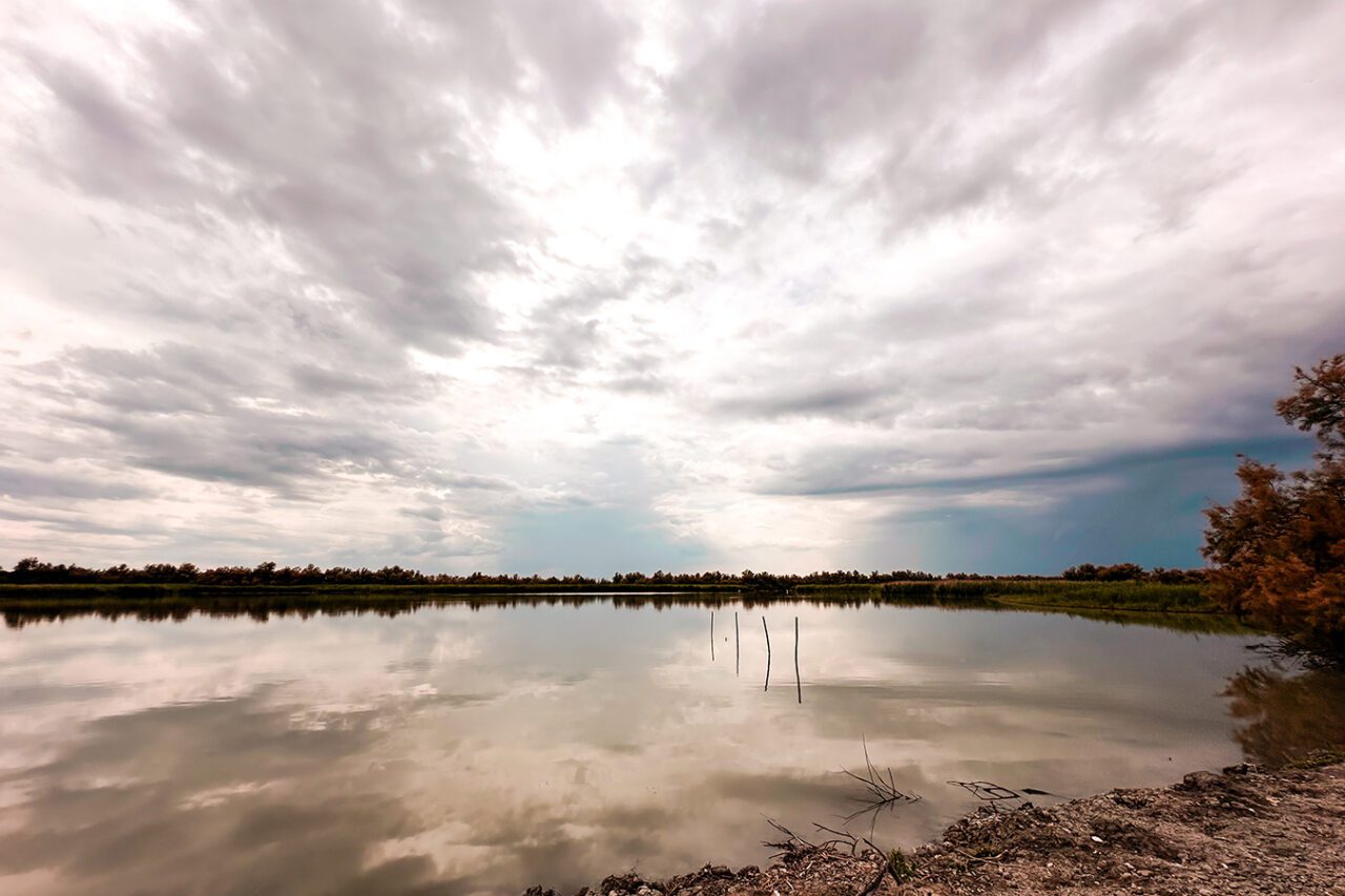 UnViaggioPerDue: Un lago calmo a Boscoforte riflette un cielo nuvoloso, circondato da alberi distanti e una costa rocciosa, incarnando la serena bellezza della Provincia di Ferrara.