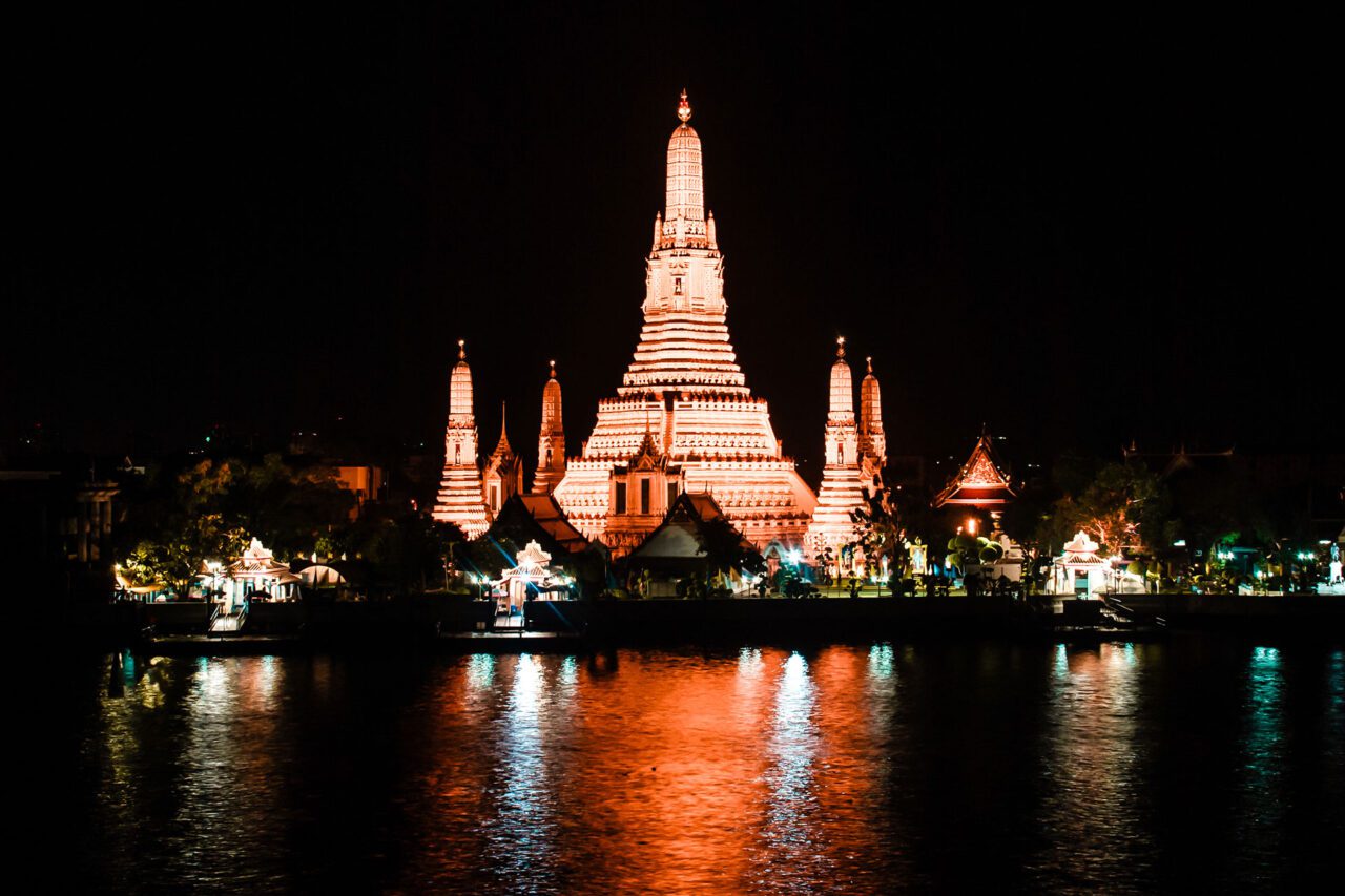 UnViaggioPerDue: Il tempio illuminato di Wat Arun si staglia contro il cielo notturno, riflettendosi brillantemente sul fiume Chao Phraya a Bangkok. Le acque serene del fiume Chao Phraya rispecchiano il suo maestoso splendore, creando un'affascinante vista notturna.