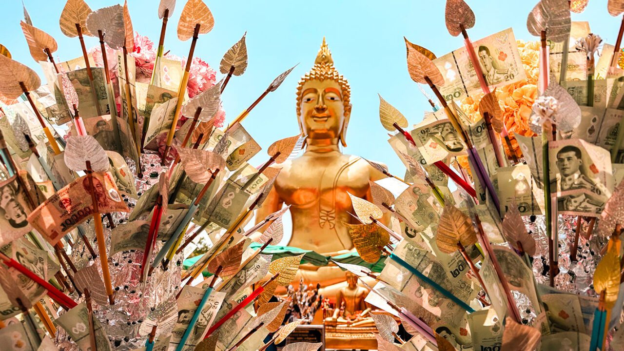 UnViaggioPerDue: una statua dorata del Buddha in Thailandia è circondata da diverse offerte monetarie sotto forma di banconote attaccate ad alberi artificiali. Le banconote sono disponibili in vari tagli e colori, aggiungendo un tocco di vivacità all'ambiente.