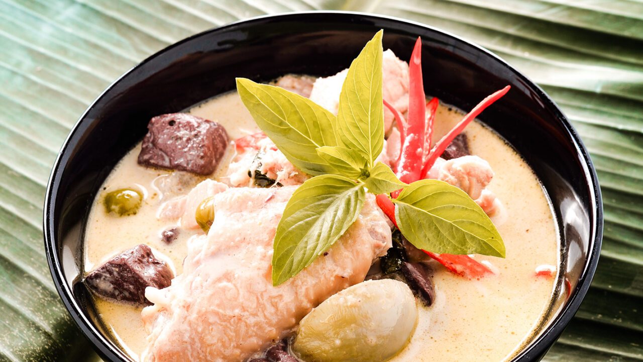 UnViaggioPerDue: una ciotola nera piena di cremoso curry verde contiene pezzi di carne di pollo e verdure, guarnita con foglie di basilico fresco e peperoncino rosso su uno sfondo di foglie verdi, catturando l'essenza della Thailandia.