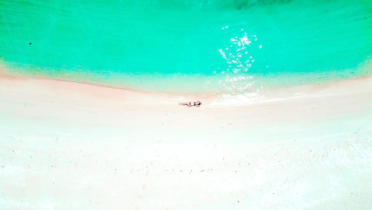 UnViaggioPerDue: Vista aerea di una spiaggia incontaminata a Koh Kham, Thailandia, con sabbia bianca, acqua turchese e cristallina e Flavia sdraiata vicino alla riva.
