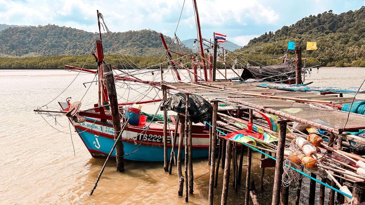 UnViaggioPerDue: una barca da pesca in legno attraccata a un molo rustico con montagne e colline boscose sullo sfondo sotto un cielo nuvoloso. Ecco la serena bellezza del villaggio dei pescatori di Koh Chang.