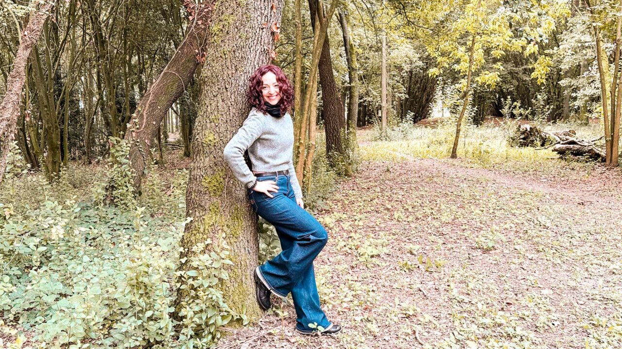 UnViaggioPerDue: Flavia si appoggia a un albero in un bosco tranquilla vicino all'Agriturismo Le Occare, indossando un maglione grigio e jeans blu. Il terreno è coperto di foglie e gli alberi circondano pacificamente la zona.